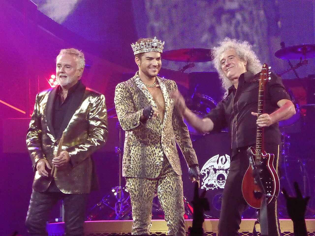Adam Lambert performing on stage with Queen in 2014, wearing a leopard-print suit and crown, flanked by band members.