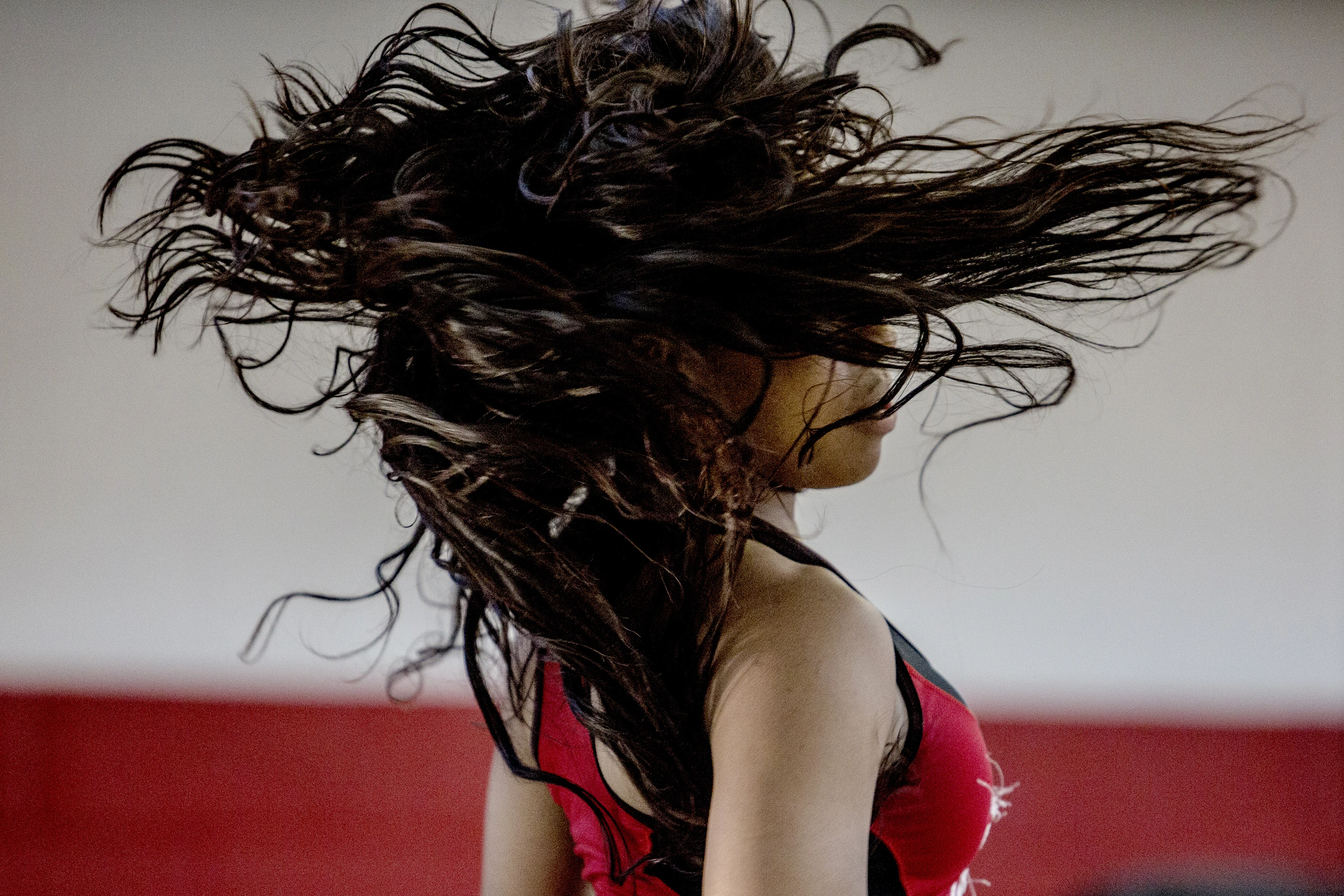 A dancer in a red outfit mid-motion with hair flying across her face against a neutral indoor background.