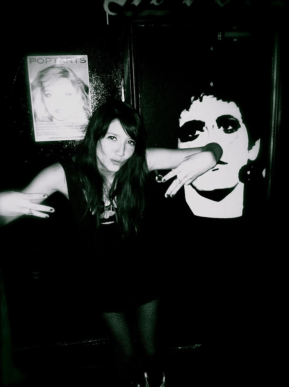 Queen Kwong posing in a dark, high-contrast candid photo with a postered wall behind her