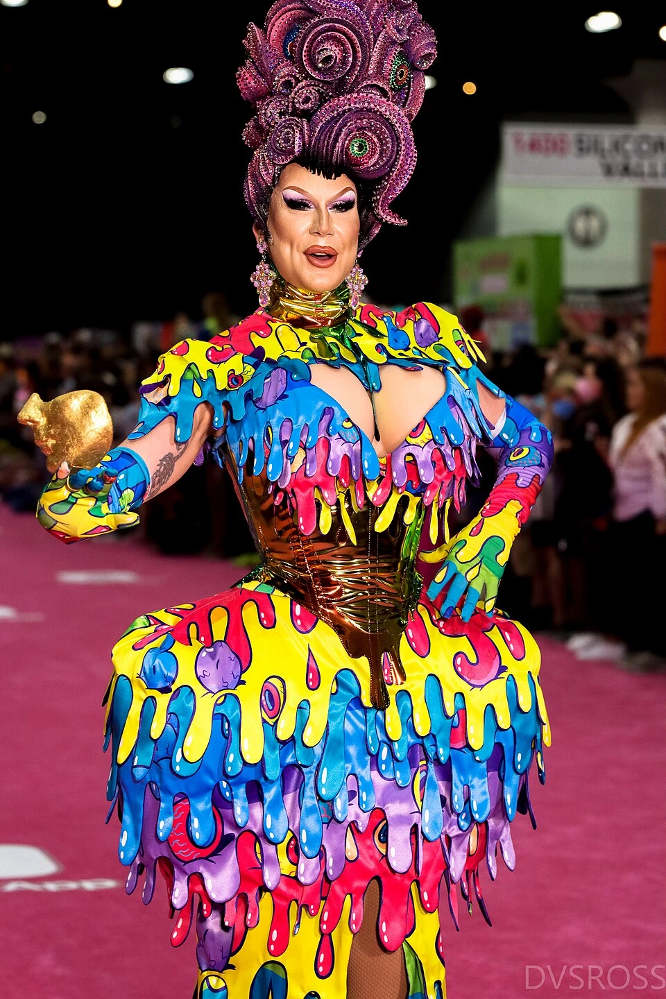 Drag queen in a colorful rainbow costume and tall floral headdress posing at a parade with other performers in front of
